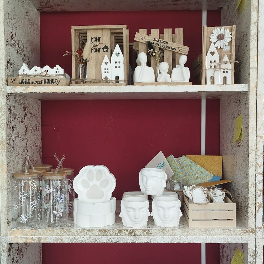 Ein weißer Regalboden zeigt Keramikartikel, einschließlich eines herzförmigen Behälters und eines Pfotenabdrucks. Unten gibt es Gläser und Boxen mit verschiedenen Designs, wie einem Blumenmuster und einem Schmetterling. Die Regale haben einen roten Hintergrund.