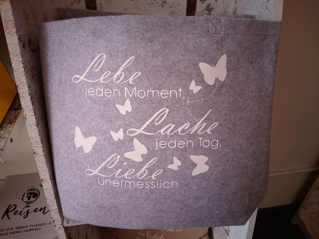 Ein grauer Tragetasche mit weißem Text 'Lebe jeden Moment, Lache jeden Tag, Liebe unermesslich' und weißen Schmetterlingsdesigns.