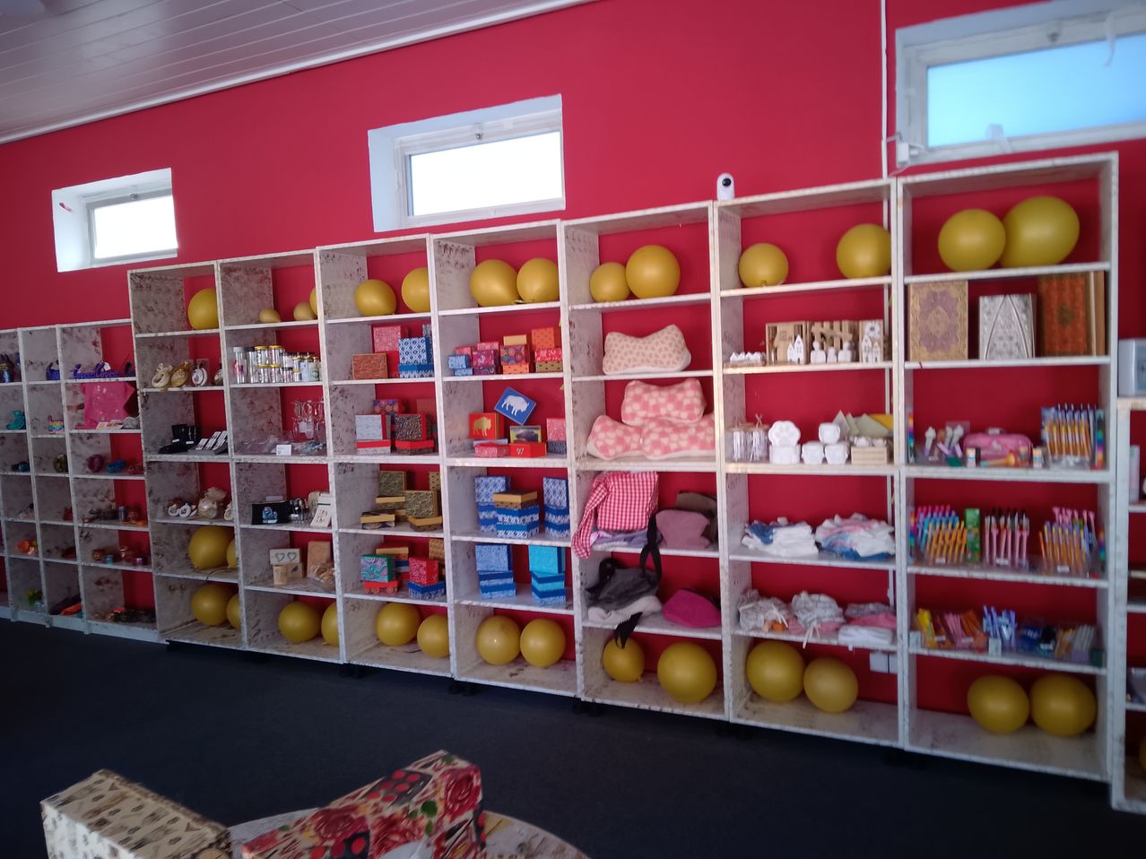 Ein rot gestrichener Raum mit Regalen, die verschiedene Gegenstände enthalten. Die Regale sind mit gelben und blauen Bällen, eingewickelten Geschenken, Kissen und anderen Objekten gefüllt. Der Raum hat zwei Fenster mit weißen Rahmen und eine weiße Decke.