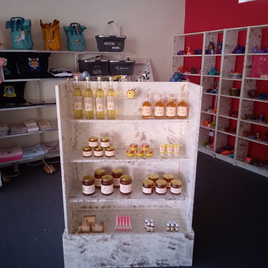 Ein Laden mit einem Ausstellungsregal, das Honiggläser und andere Produkte enthält. Regale mit Taschen und verschiedene Artikel sind ebenfalls sichtbar.