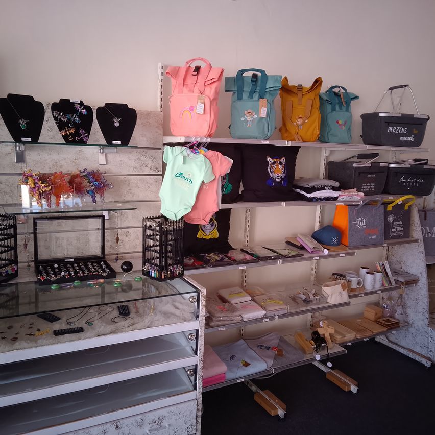 Ein Laden zeigt Taschen, Kleidung und Schmuck auf Regalen. Mannequins halten Halsketten. Ein Glaskasten enthält Schmuck. Ein Regal hat Taschen, Tassen und einen Korb mit der Aufschrift Herzens.