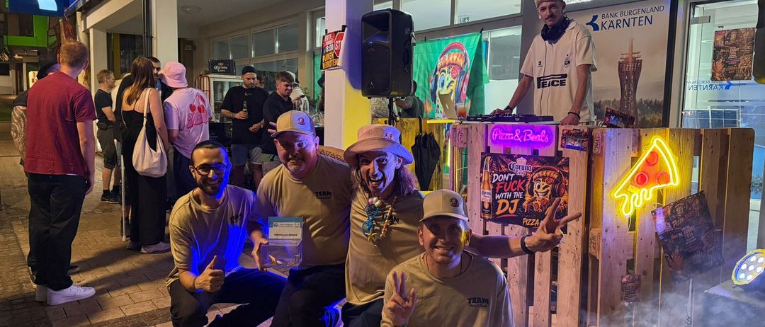 Vier Männer posieren für ein Foto vor einem DJ-Pult mit Neon-Schildern und einer Menschenmenge im Hintergrund.