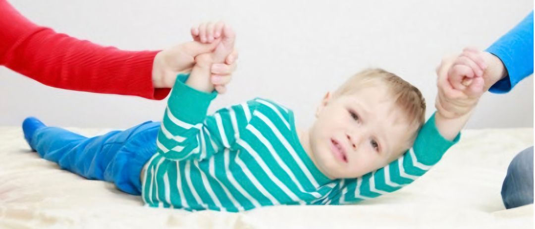 Ein Baby in einem blau-weiß gestreiften Shirt liegt auf einem Bett und wird von einer Person in einem roten Langarmshirt gehalten.