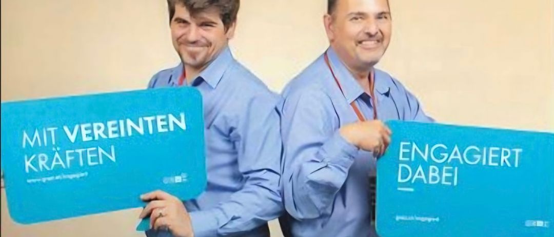 Zwei Männer in blauen Hemden halten Schilder mit den Aufschriften 'Vereinten' und 'Engagieren' und lächeln für ein Foto.