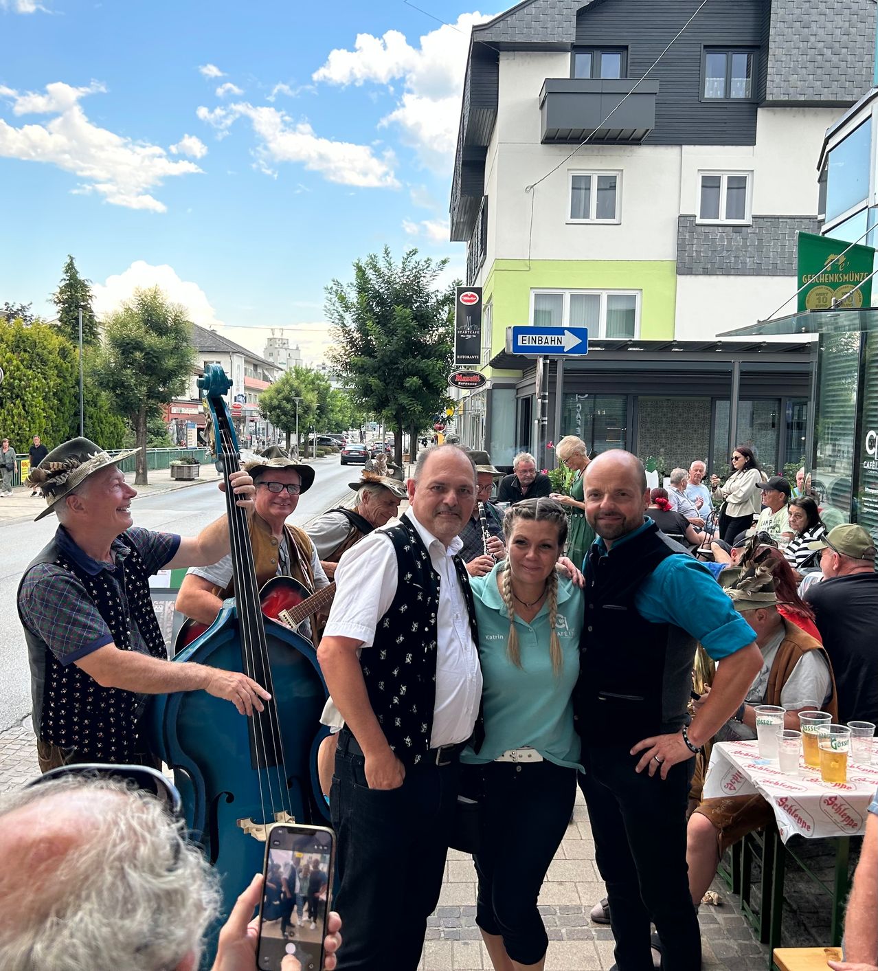 A group of people, including a man playing a bass guitar, are standing on a sidewalk in front of a building with a sign that reads 'Einbahn'. They are smiling and posing for a photo. There are tables with glasses of beer and people sitting on chairs in front of the building. Trees and a car are visible in the background.