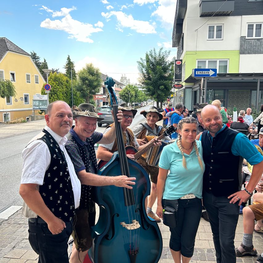 A group of musicians in traditional attire pose for a photo on a sunny street with buildings and trees in the background.