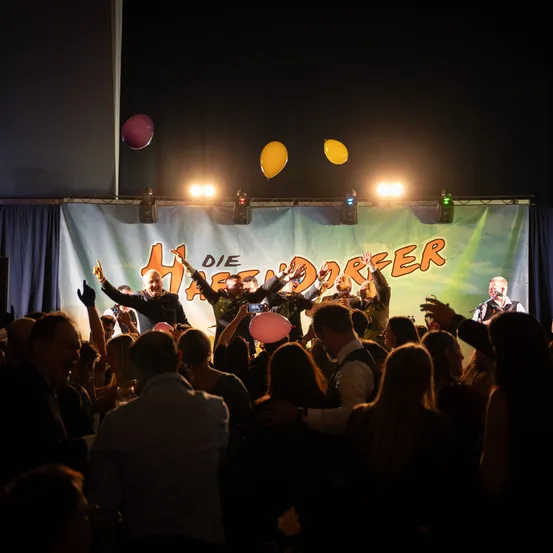 Eine Menschenmenge feiert bei Nacht auf der Bühne, mit Ballons und einem Banner mit der Aufschrift 'DIE HAGENDORFER'. Darsteller und Gäste sind in bester Stimmung.