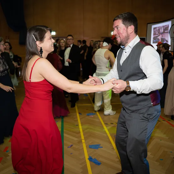 Ein Mann und eine Frau tanzen zusammen in einem Raum. Die Frau trägt ein rotes Kleid und der Mann trägt eine Weste. Es gibt andere Leute im Raum, einige beobachten sie. An der Wand befindet sich eine Projektionsleinwand.