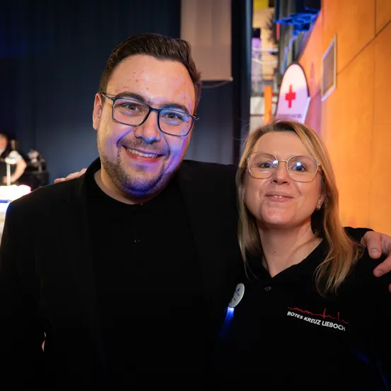 Ein Mann und eine Frau stehen lächelnd eng beieinander vor einem Gebäude mit einem roten Kreuz-Symbol. Der Mann trägt eine Brille und ein schwarzes Hemd, während die Frau ein Licht auf ihrem Shirt hat.