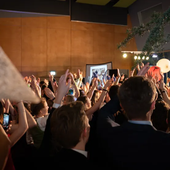 Eine Menschenmenge, die die Hände hebt, wahrscheinlich bei einer Konferenz oder Veranstaltung. Einige machen Fotos mit Handys. Der Raum hat Holzwände und eine Projektionsleinwand.