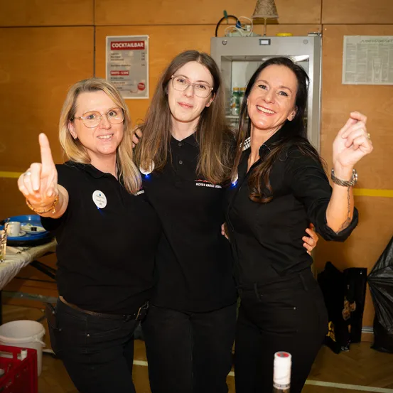 Drei Frauen in schwarzen Uniformen stehen zusammen, zwei mit Armen umeinander und eine mit erhobenem Daumen. Sie befinden sich in einem Raum mit einer Holzwand und einem Cocktailbar-Schild.