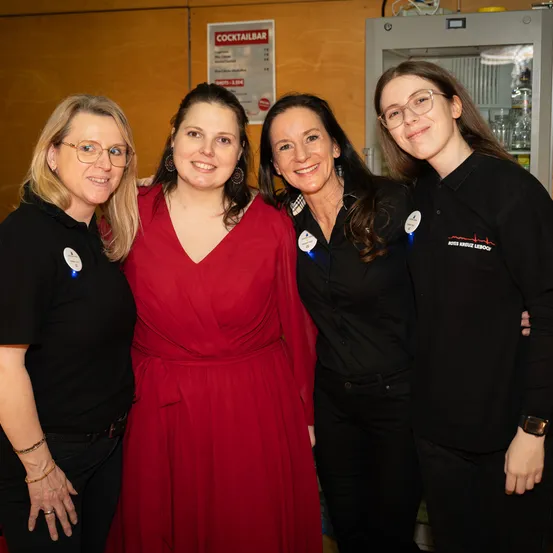 Vier Frauen in schwarzen Shirts stehen in einem Raum zusammen. Eine trägt ein rotes Kleid und hat den Arm um eine andere Frau gelegt. Sie alle haben Namensschilder. Hinter ihnen befindet sich eine Holzwand mit einem Schild für eine Cocktailbar.