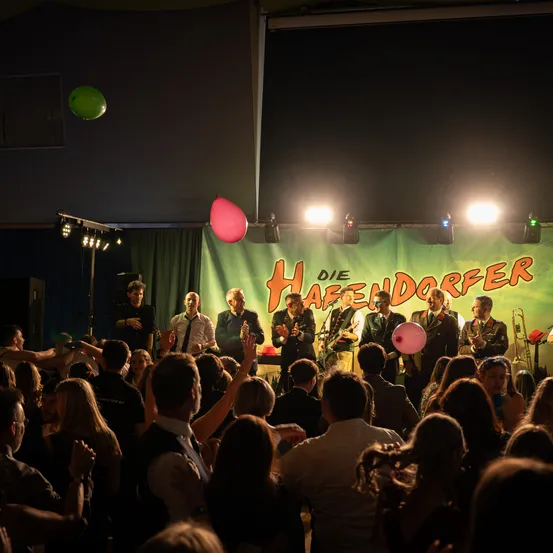 Eine Band tritt auf einer Bühne mit einem Banner auf, das Die Hasendorper sagt. Das Publikum jubelt.