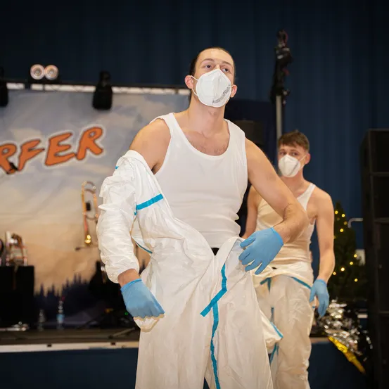 Zwei Männer auf der Bühne, beide tragen Masken und Handschuhe, einer justiert seinen Schutzanzug, während ein anderer hinter ihm steht. Ein Banner mit der Aufschrift 'Surfer' und einer landschaftlichen Kulisse im Hintergrund.