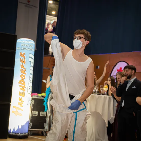 Ein maskierter Mann in einer weißen Tunika und Handschuhen tritt auf der Bühne auf. Hinter ihm klatschen Menschen. Ein Banner mit der Aufschrift Hafendorfer ist zu sehen. Ein Tisch mit Weingläsern steht im Hintergrund.