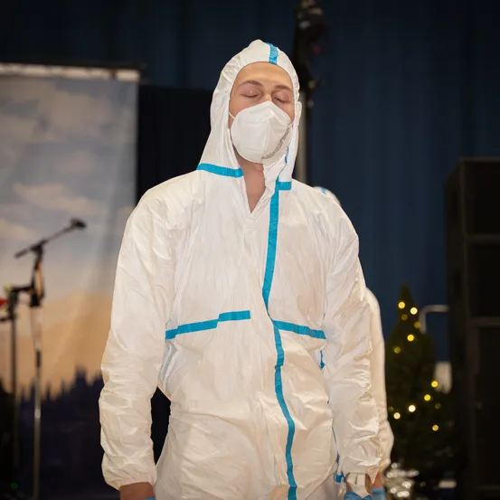 Eine Person in einem weißen Schutzanzug und Maske steht auf der Bühne, Augen geschlossen, mit einem Mikrofon und einem Weihnachtsbaum im Hintergrund.