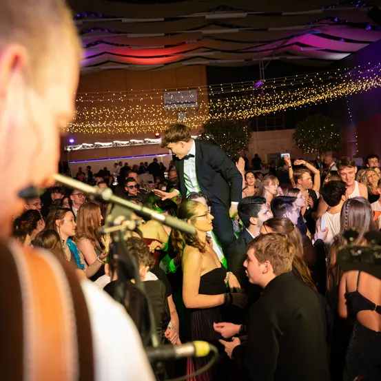 Eine große Menschenmenge auf einer Party, mit einem Mann auf den Schultern einer Frau. Lichter und Bäume schmücken den Veranstaltungsort.