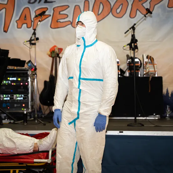 Eine Person in einem Schutzanzug steht auf einer Bühne mit einem Banner mit der Aufschrift Hafendorf hinter ihr. Sie trägt eine Maske und Handschuhe.