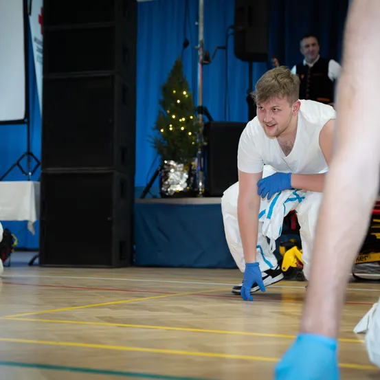 Ein Mann in Schutzkleidung kniet lächelnd auf einem Basketballplatz, mit einem Weihnachtsbaum und einem Lautsprecher im Hintergrund.