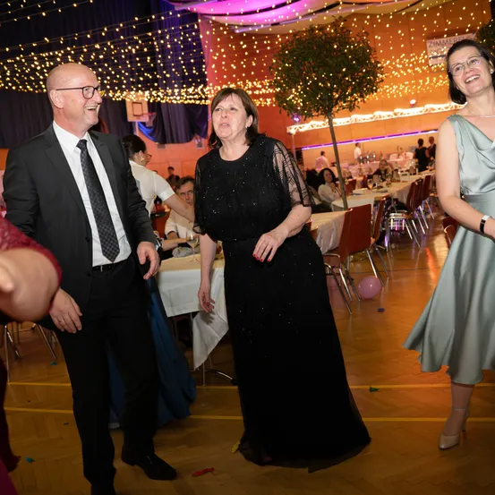 Ein Mann und zwei Frauen stehen in einem Bankettsaal. Die Frau in der Mitte trägt ein schwarzes Kleid. Hinter ihnen sind Tische für ein Bankett gedeckt, an denen Leute sitzen.