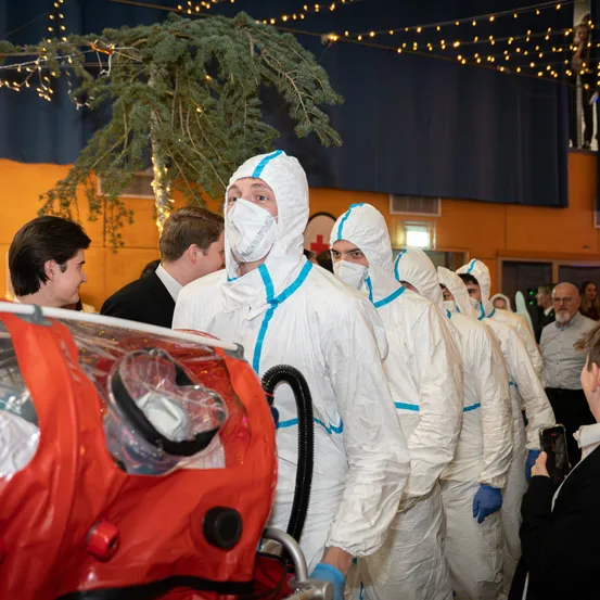 Eine Gruppe von Menschen in Schutzanzügen steht neben einem roten medizinischen Container. Sie sind drinnen, mit einem Weihnachtsbaum und festlichen Lichtern im Hintergrund.