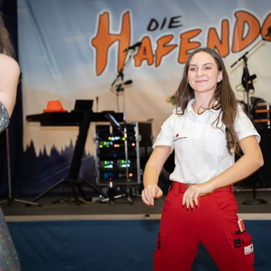 Eine lächelnde Frau in roten Hosen und einem weißen Shirt steht auf der Bühne, wahrscheinlich bei einer Aufführung. Hinter ihr befindet sich ein Hintergrund mit dem Wort 'HAFEN' und einem Banner mit Musikinstrumenten.