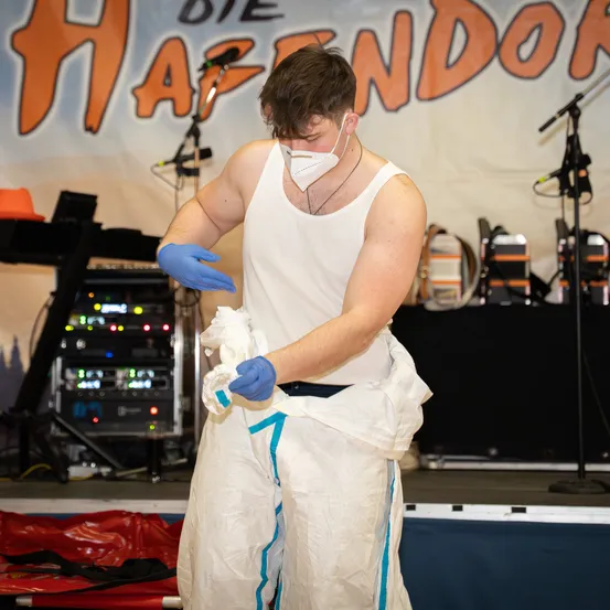 Ein Mann in einem weißen ärmellosen Shirt, Gesichtsmaske und blauen Handschuhen steht auf einer Bühne mit einem Banner, das 'Haendor' im Hintergrund steht.