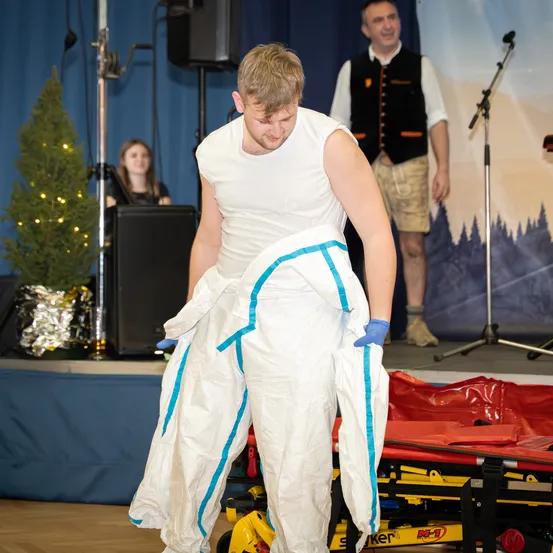 Ein Mann in einem weißen Schutzanzug und Handschuhen geht auf der Bühne, während ein anderer Mann in einer Weste hinter ihm steht. Ein Weihnachtsbaum und ein Lautsprecher befinden sich auf der linken Seite der Bühne.