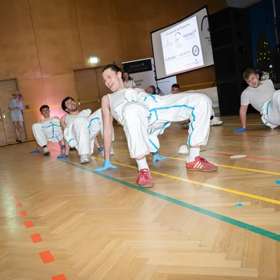 Eine Gruppe von Männern, die in weißes Kleidungsstück mit blauen Streifen gekleidet sind, führt eine Übungsroutine auf einem Turnhallenboden mit Markierungen durch. Ein Beamerbildschirm ist im Hintergrund zu sehen.