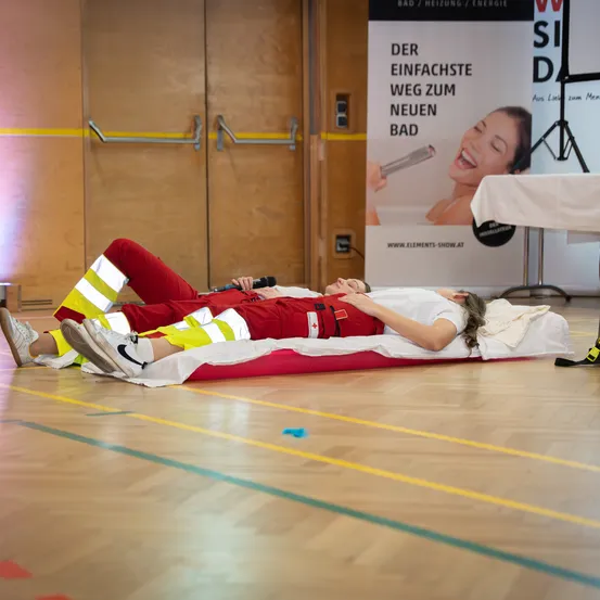 Zwei Personen liegen auf einer roten Matratze in einer Turnhalle, in Schutzanzügen. Eine hält ein Mikrofon, und eine Werbung ist im Hintergrund sichtbar.