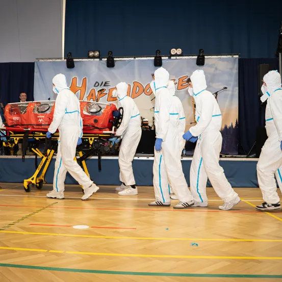 Eine Gruppe von Personen in weißen Schutzanzügen trägt eine rote Trage auf einer Bühne, wahrscheinlich für eine Aufführung oder Simulation.