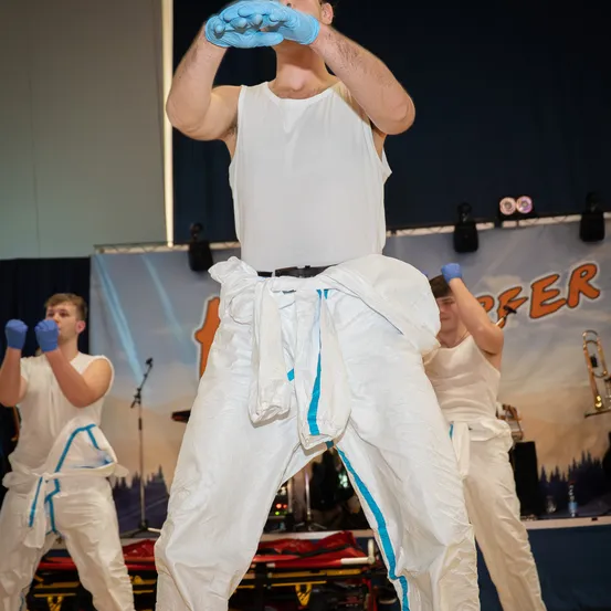 Ein Mann in einem weißen Tanktop und weißen Hosen mit blauen Streifen tritt auf der Bühne auf. Zwei weitere Personen treten ebenfalls hinter ihm auf. Sie tragen alle Handschuhe und es gibt ein Banner hinter ihnen.