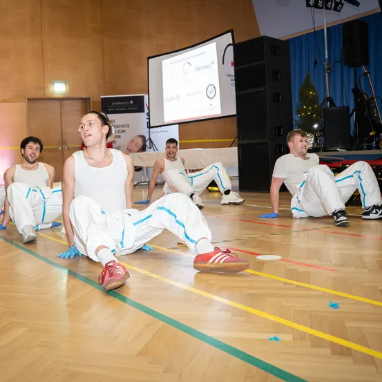 Fünf Männer in weißen Outfits sitzen mit Turnschuhen und blauen Handschuhen auf dem Boden in einem Raum mit Beamer, Tisch und Weihnachtsbaum.