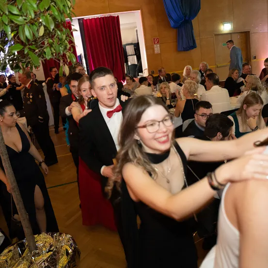 Eine lebhafte Indoor-Veranstaltung mit tanzenden und sozialisierenden Menschen, darunter ein Mann im Smoking und eine Frau mit Brille.