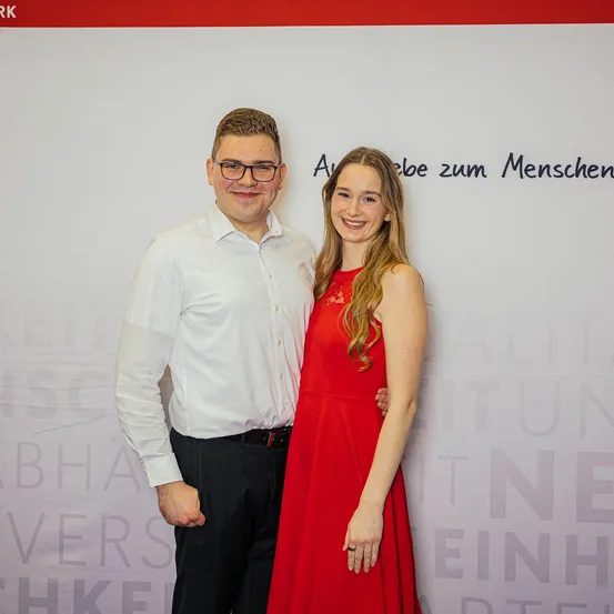 Ein Mann und eine Frau stehen vor einem weißen Hintergrund mit Text und lächeln für ein Foto. Der Mann trägt eine Brille und ein weißes Hemd, während die Frau ein rotes Kleid trägt.