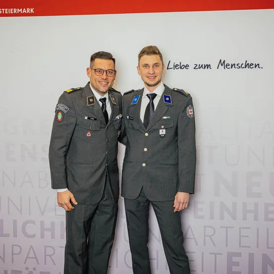 Zwei Männer in Militäruniformen stehen nebeneinander und lächeln für ein Foto. Der Hintergrund zeigt eine große weiße Banner mit Text.