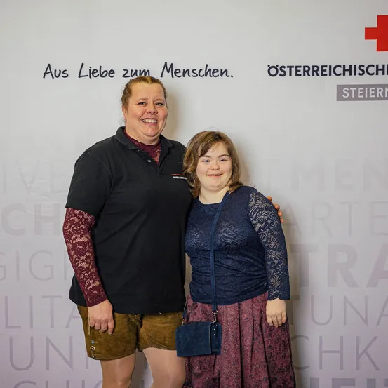 Zwei Frauen stehen lächelnd eng beieinander vor einer weißen Wand mit Text und rotem Kreuz. Die Frau auf der linken Seite trägt ein schwarzes Shirt und braune Shorts, während die Frau auf der rechten Seite eine blaue Bluse und einen kastanienbraunen Rock trägt.