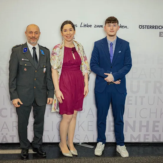 Drei Personen stehen zusammen und posieren für ein Foto. Der Mann links trägt eine Militäruniform. Die Frau in der Mitte ist in einem Purpurkleid gekleidet. Der Mann rechts trägt einen blauen Anzug. Hinter ihnen ist eine weiße Wand mit Text.