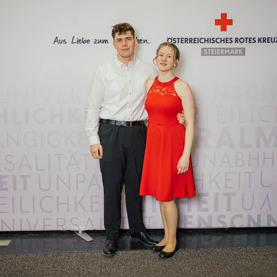 Ein Mann und eine Frau posieren in formeller Kleidung für ein Foto vor einer Wand mit dem Roten Kreuz-Logo und Text.