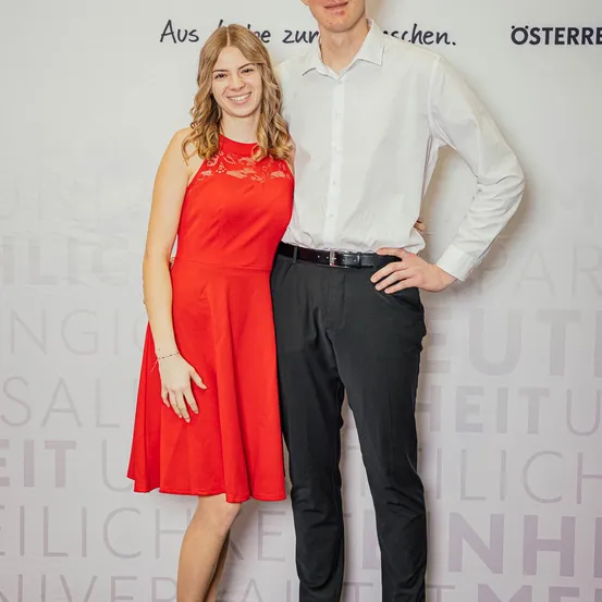 Ein Mann und eine Frau stehen nah beieinander und lächeln. Die Frau trägt ein rotes Kleid, der Mann ein weißes Hemd und schwarze Hose. Sie posieren vor einem weißen Hintergrund mit verschwommenem Text.