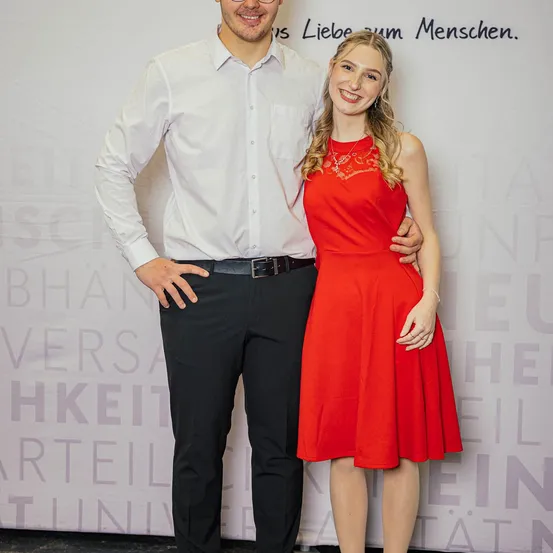 Ein Mann und eine Frau stehen lächelnd nebeneinander. Der Mann trägt ein weißes Hemd, schwarze Hose und einen Gürtel. Die Frau trägt ein rotes Kleid. Hinter ihnen ist eine weiße Wand mit Text.