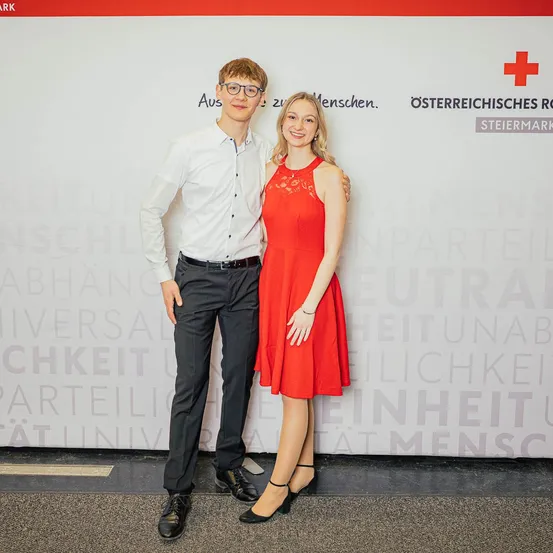 Ein junger Mann und eine junge Frau stehen eng beieinander vor einer Wand mit verschiedenen aufgeschriebenen Worten, darunter 'Aus zwei Menschen' und 'Osterreichisches Rot Kreuz'.