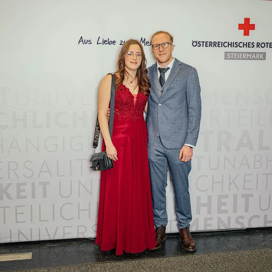 Ein Mann und eine Frau in formeller Kleidung stehen nebeneinander vor einer Wand mit Wörtern und einem Logo.