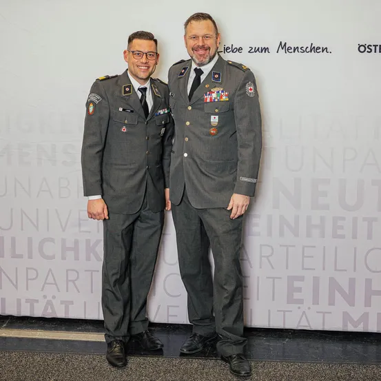Zwei Männer in Militäruniformen, einer mit Brille, stehen zusammen vor einer weißen Wand mit deutschem Text und Logos.