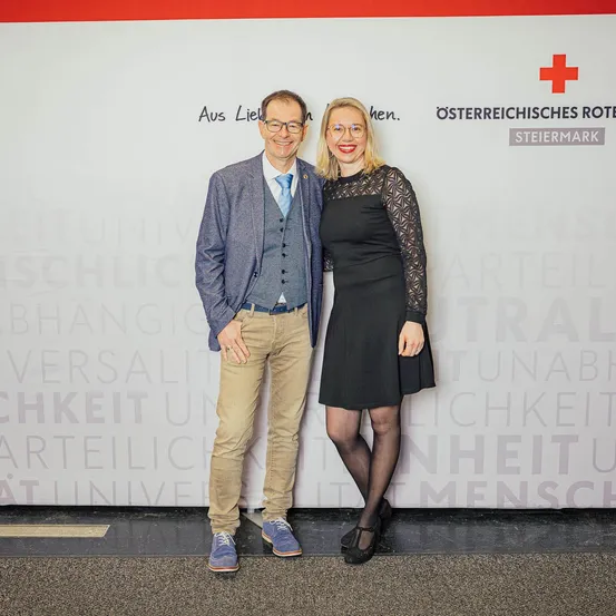 Ein Mann und eine Frau stehen nebeneinander und lächeln für ein Foto. Der Mann trägt eine Brille, eine Krawatte und Schuhe. Die Frau trägt ein schwarzes Kleid und eine Brille. Hinter ihnen ist eine Wand mit Text und einem roten Kreuzlogo.