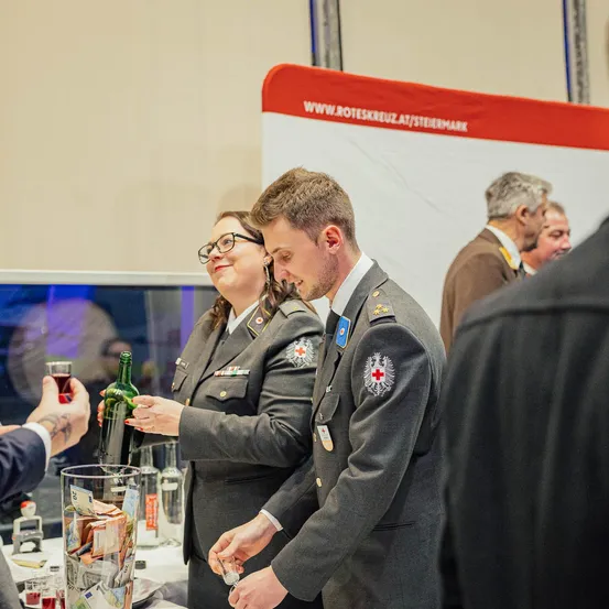 Zwei Personen in Militäruniformen bereiten sich darauf vor, eine Flasche bei einer Veranstaltung zu öffnen. Ein Banner mit einer Website ist im Hintergrund sichtbar.
