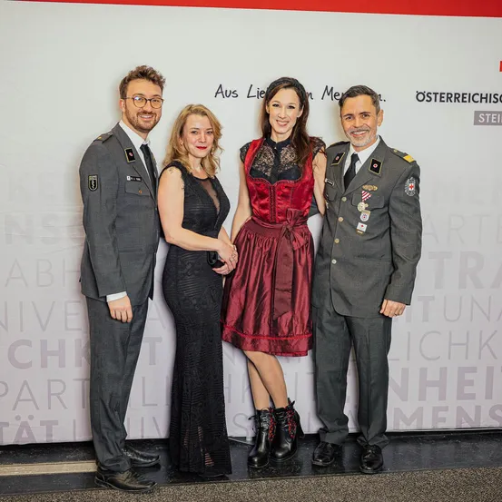 Vier Menschen stehen vor einer Wand mit der Aufschrift 'Aus Liebe zu Men'. Die beiden Männer sind in Uniform. Die beiden Frauen lächeln. Die Frau rechts trägt ein rotes Kleid und Stiefel.