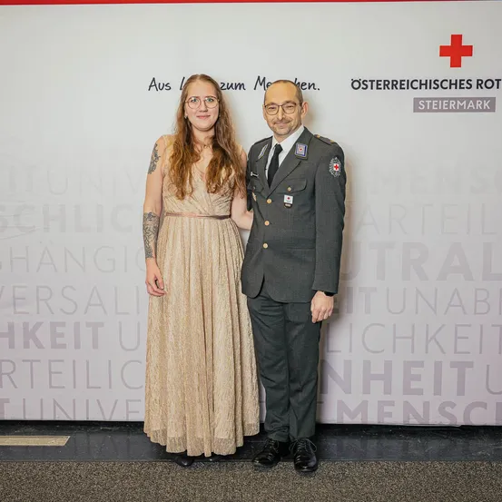 Ein Mann und eine Frau in formeller Kleidung stehen nah beieinander und lächeln. Der Mann trägt eine Militäruniform mit einem roten Kreuz auf der Brust. Sie stehen vor einer Wand mit Text, der 'Osterreichisches Rot Steiermark' lautet.