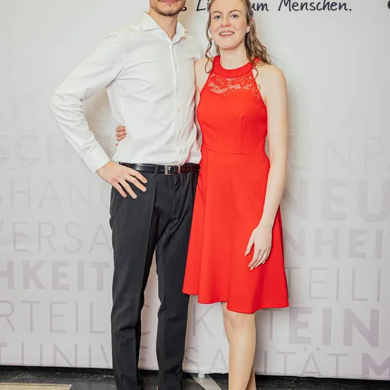Ein Mann und eine Frau stehen gemeinsam in formeller Kleidung und lächeln in die Kamera. Der Mann trägt ein weißes Hemd und schwarze Hose, und die Frau ist in einem roten Kleid. Sie stehen vor einer weißen Wand mit darauf geschriebenen Worten.
