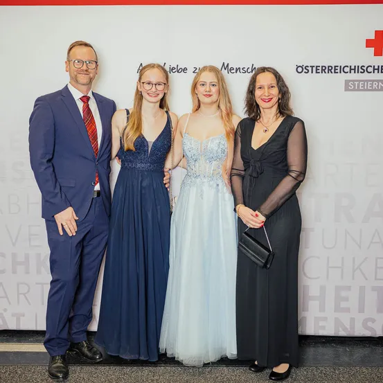 Vier Personen stehen zusammen, wahrscheinlich für ein Foto, mit einer Wand im Hintergrund, die Text und ein rotes Kreuz-Logo aufweist.
